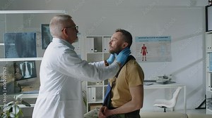 Middle-aged male doctor in lab coat and medical gloves palpating lymph nodes of his soldier patient in military uniform with broken arm, sitting on examination couch in clinic Stock Video