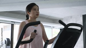 45 degrees front view, low angle medium shot portrait of of long hair middle-aged Asian woman, exercising, using elliptical machine, workout alone at fitness gym. Sport, health, wellbeing, concept Stock Video