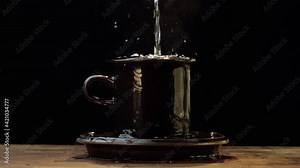 Cup overflows onto the saucer and table. Slow motion. Dark studio setting.