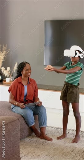 Vertical video: African American mom holding tablet watching child in VR reaching for items at home
