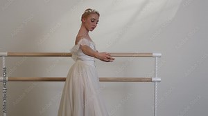 A beautiful ballerina in a white dress dances in a white sunny room with a black floor next to a ballet barre.
