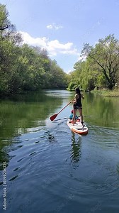 A woman paddling on a stand-up paddleboard with a child seated, navigating a calm river surrounded by lush green trees under a bright blue sky