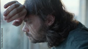 Depressed man staying alone near window with stressed problem face, closed eyes. Pensive, thoughtful. Denial, Bargaining emotions. Mental health, man depression, despair concept. Long hair, beard