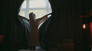 Close up back view young woman opening window curtains in luxury apartment home or in spa hotel looking through window enjoying mountain landscape. Warm morning light. Woman on summer vacation.