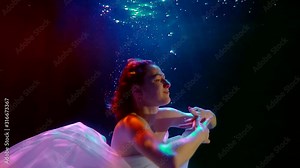 a girl with long hair and a white dress floats under the water in the dark. she moves her hands beautifully, spreads them out to the sides and floats up. blue and red backlight