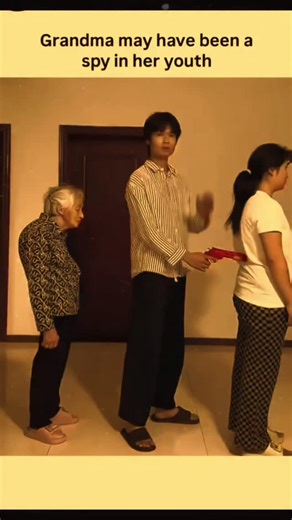 Explainer Zone™ on Instagram: "The video takes place in a simple indoor room with white walls, a brown door, and tiled floors, featuring an elderly woman with short gray hair wearing a floral-patterned top and black pants, standing alongside a young man in a striped shirt and black pants, and a young woman in a white t-shirt and checkered pants observing. It begins with the young man playfully holding a red toy gun to the back of the young woman's head, demonstrating a mock threat, but she strug