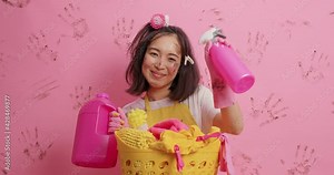 Glad Asian housewife shows bottle of liquid washing powder and spray detergent does laundry at home has cheerful expression after finishing work poses against background. Housekeeping cleaning concept