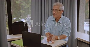 Older male professor teaching remotely from classroom, engaging in online learning using laptop and digital pen.