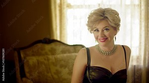 Attractive woman wearing a classic fifties dress and sitting on a chaise lounge looks at camera and smiles. Medium shot, recorded in 4K and sepia tinted.
