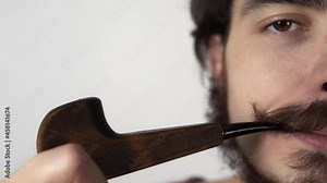 Closeup footage of young man with curly mustache smoking pipe standing on white background. Isolated portrait of a bearded male person fuming tobacco and exhaling steam. Young hipster with retro tube.