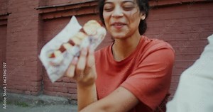 A young woman eats a hot dog and smiles while sitting on a longboard next to a man on a city street