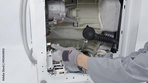 A repairman is carefully dismantling a washing machine. He is using a screwdriver, a drill and other tools.