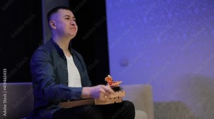 Man using tv remote for changing channel while eating pizza. Focused young man is eating pizza while watching tv sitting in the kitchen at home.