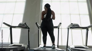 The back side view of Body confident young woman dancing in sportswear while standing on a treadmill near panoramic window in modern gym. Sun shining through window.