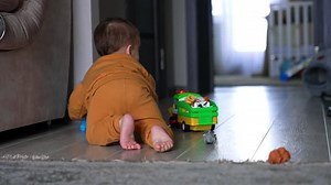 Toy car is moving by the floor and barefoot baby boy follows it. Kid sits on his knees and presses the button on a toy. Rear view.