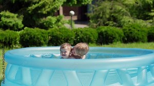 Little siblings having fun in inflatable swimming pool in backyard in slow motion. Girl embracing and kissing her cute toddler brother laughing joyfully on summer vacation day Stock Video