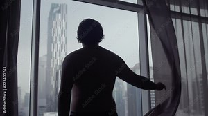 Fat man open curtains at midday, stay against window and look to urban city view from high floor. Black silhouette, half length view from back scratching his ass. slow motion. 3840x2160