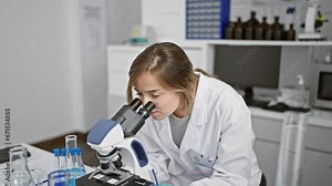 Confident young blonde woman scientist, smiling with crossed arms, sitting by the lab table, working on medical research with microscope at the bustling science laboratory.
