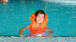 A child in an orange life jacket swims in the pool in an aqua complex for family holidays. Safety on the water, learning to swim