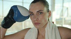 Close-up portrait of beautiful young woman fighter wearing boxing gloves and towel standing in gym alone and looking at camera. People and sports concept.