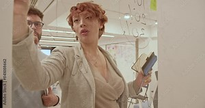 Two colleagues collaborate on a new project, writing ideas, details and sharing knowledge on a glass wall in a bright office environment.