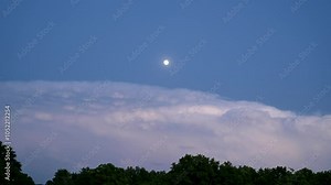 Time lapse of thunderstorm clouds with lightning moving to the left above trees, with a full moon rising above the clouds, lighting them from above