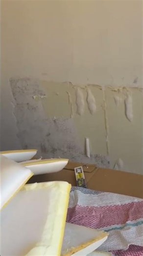 applying cream colored adhesive paste onto a beige indoor wall using a small metal putty knife tool