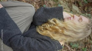 Blonde girl is relaxing on the ground in a peaceful landscape, enjoying the beautiful landscape with trees and plants surrounding her. She feels the soil beneath her feet. Vertical video