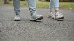 Lower angle view of two people walking side by side in grey joggers and sneakers on paved road with visible motion, relaxed stride, and outdoor setting surrounded by greenery and blurred elements | Premium Stock Video Footage
