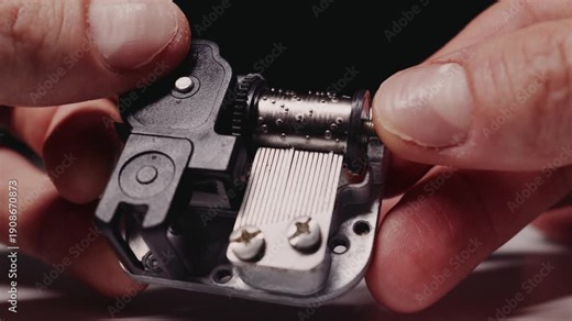 Fingers wind the gears of a metal music box mechanism causing the cylinder to rotate and play a tune. The pins pluck the comb against a black background. Concept of mechanical precision and nostalgia