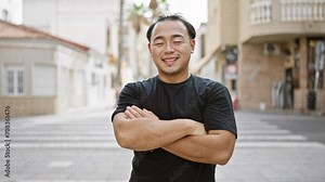 Cheerful young chinese man, radiating confidence and joy, spotted smiling and standing with crossed arms gesture on a bustling urban street