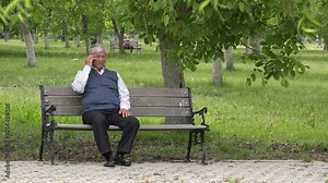 Single old man sits on bench talking at mobile phone in park