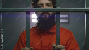 Male criminal in orange uniform and handcuffs holds metal bars, stands in prison cell, looks at camera. Prisoner serves imprisonment term in jail. Detention center or correctional facility. Portrait.