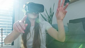 Young woman wearing virtual reality glasses, exploring the augmented world, interacting with the digital interface while sitting in the living room, handheld shot.