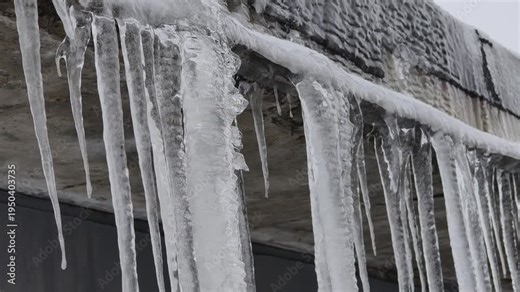 Thick, clear melting icicles hang from a concrete roof edge during freezing winter weather. Concept of extreme cold, seasonal climate shifts, and dangerous freezing weather hazards.