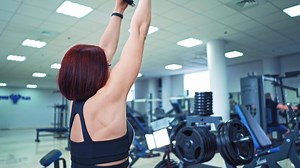 Beautiful strong woman bodybuilder is doing workout to build her muscles in sports gym. Muscular red-haired girl in sportswear exercising heavily on a modern simulator.