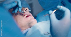 Close-up an adult male patient is under anesthesia while the dentist is holding a dental retractor and pulling forceps with the surgical thread and a nurse is inserting a saliva ejector.