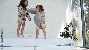 Jumping with pillow on the bed together. Two cute little girls in nightwear at daytime in modern room