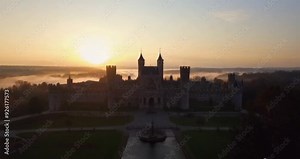 An aerial view captures a majestic castle at sunrise, surrounded by lush greenery and mist. The golden light highlights the castle’s towering architecture, creating a serene and mystical atmosphere.