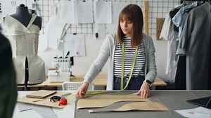 Young pretty seamstress is measuring clothing patterns with tape-measure in her modern tailors shop. Getting ready for sewing clothes concept.