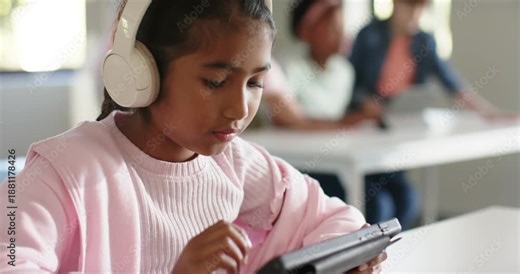 Female child reacting to tablet prompt, tapping cased tablet, wearing white headphones in classroom