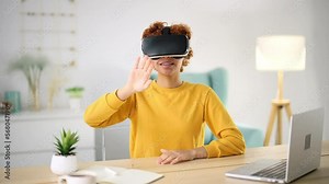 Young woman at home put 3D glasses of virtual reality, sits at desktop near laptop. Amazing girl smiles, controls video game with hand movements and gestures, touching air. New technology of future.
