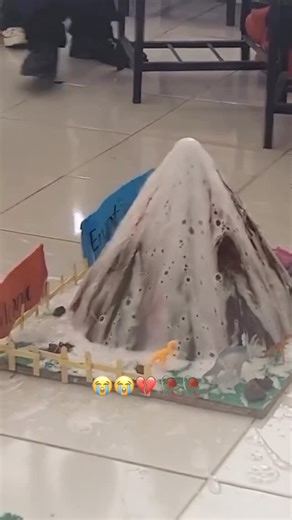 @the.reel.nigga on Instagram: "😭😭 This video captures a classroom setting where several students have presented their model volcanoes for a science project. The main focus is on a large, detailed volcano model as a student pours a liquid—likely vinegar—into the crater to trigger a chemical reaction with baking soda. Instead of a typical “lava” eruption, the reaction produces a massive, overflowing fountain of thick white foam that quickly covers the entire model and spills onto the floor. The