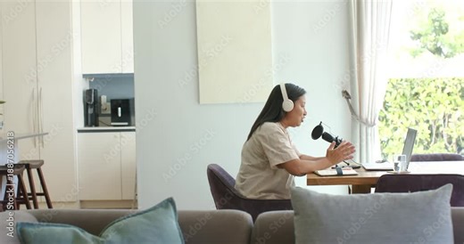 Mature Asian woman starting podcast, sitting table with headset speaking into mic checking laptop