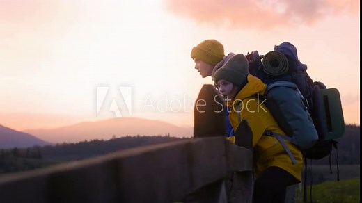 Diverse young hikers near wooden fence look at beautiful sunset and mountain scenery enjoying nature during trip. Teenage tourists rest after hike in the mountains. Outdoor enthusiasts. Slow motion.