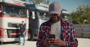 Portrait of african american lorry driver using smartphone for texting outdoors next to his trunk. Truck driver holding phone checking product list, Driver writing electronic log books