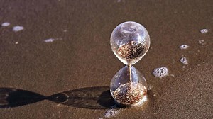 Glass sand timer stands on beach with transparent sea waves