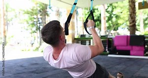 Male person uses training machine to pump up arms in light home gym. Fit man in sportswear uses exercise equipment for intensive workout slow motion