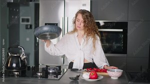 Young curly woman puts tomatoes on hot pan to fry them with scrambled eggs. Young female cooking omelette on gas stove. Girl preparing her breakfast at the modern minimalistic kitchen island.