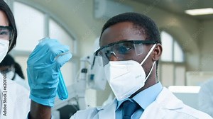 African American scientists working at laboratory room. Multiethnic woman and man dressed up in safety glasses and medical masks. Male checking test tube with blue substance.